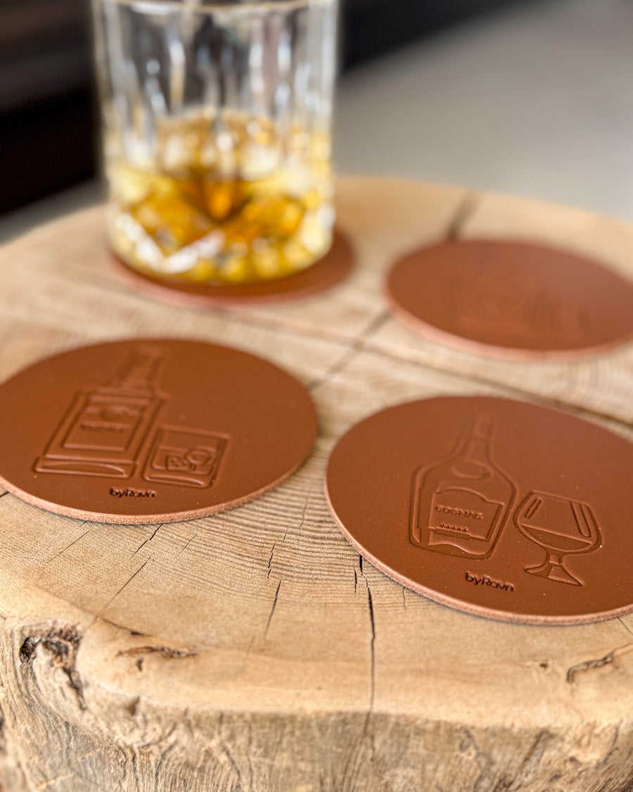 MOKKA spirits coasters in cognac leather embossed with whisky and cognac on a wooden slab by a crystal glass of whisky with ice