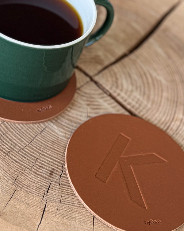 MOKKA coaster in cognac leather embossed with the letter "K" on a wooden slab next to a green coffee cup on a coaster