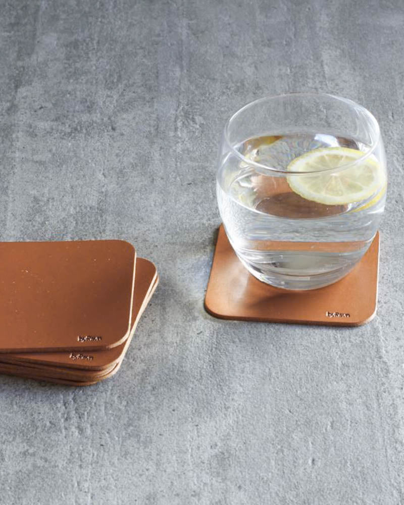 MOKKA square coasters in cognac leather stacked and under a glass of water with lemon on a concrete table