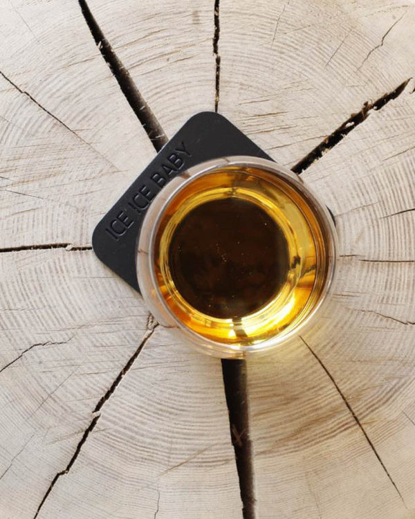 MOKKA square coaster in black leather with the engraving "Ice ice baby" under a whisky glass on a wooden slice, seen from above