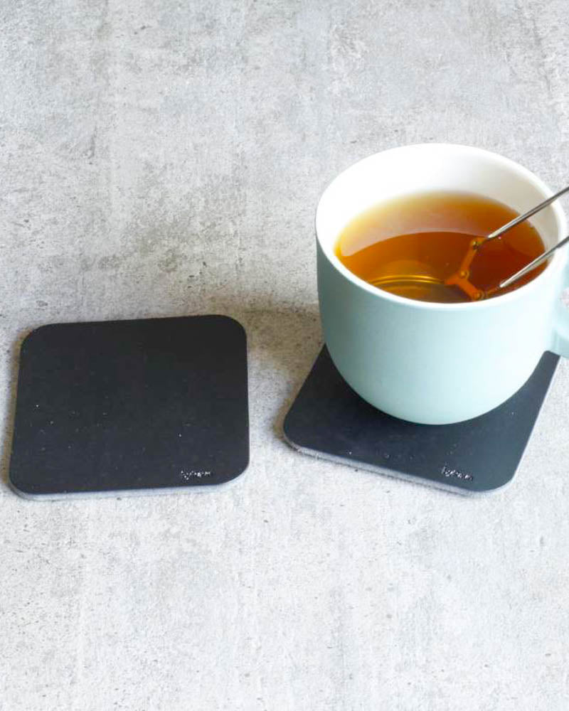 MOKKA square coasters in black leather on a concrete table, one under a light blue cup of tea