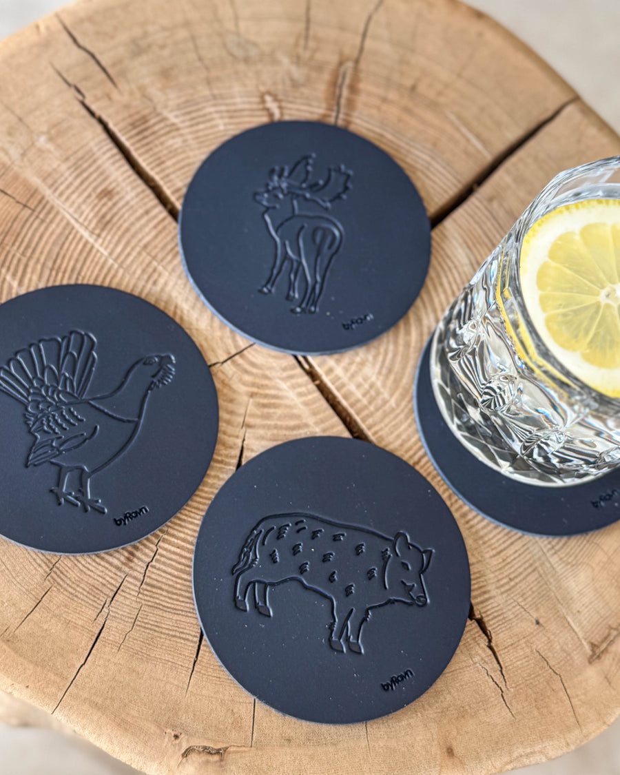 MOKKA hunting coasters in navy blue leather embossed with wild boar, capercaillie and fallow deer on a wooden slab by a glass with lemon