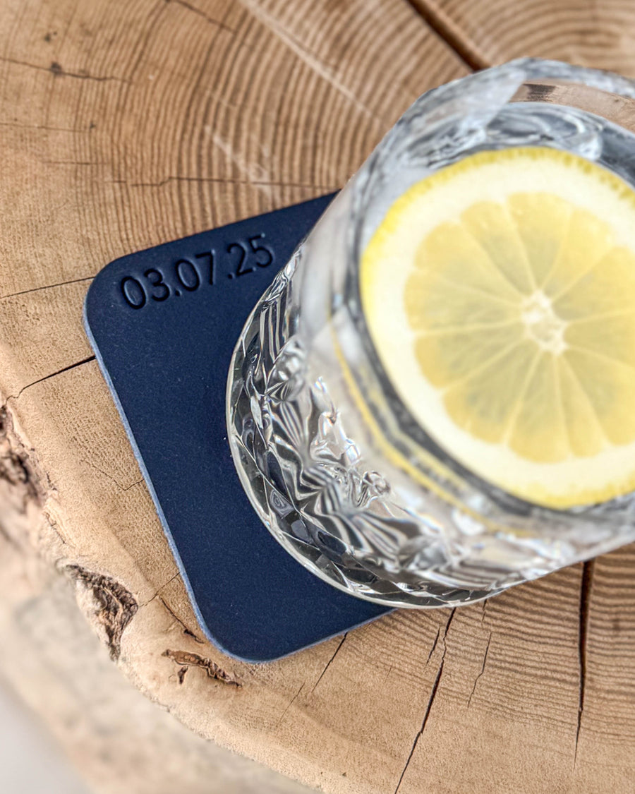 MOKKA square coaster in navy blue leather engraved with the date "03.07.25" under a crystal glass with lemon slice on a wooden slab