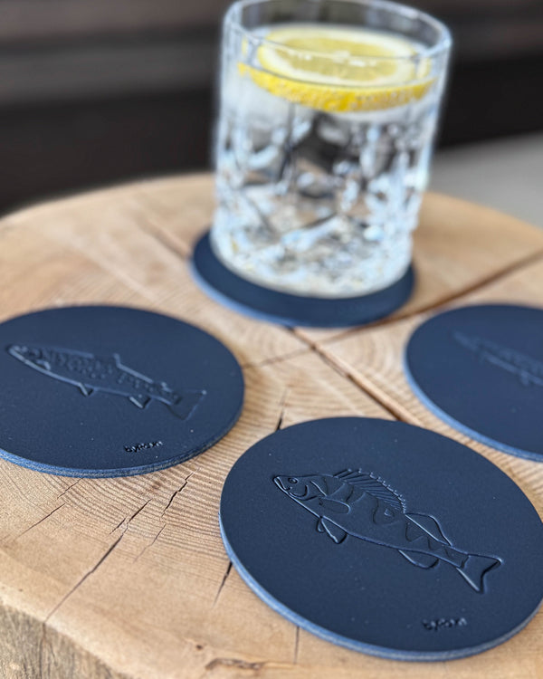 MOKKA angler coasters in navy blue leather embossed with perch and sea trout on a wooden slab by a glass with lemon slice