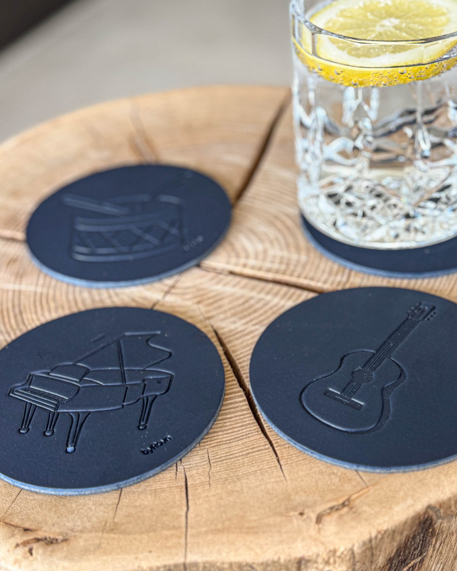 MOKKA music coasters in navy blue leather embossed with grand piano, guitar and drum on a wood slice by a glass with lemon