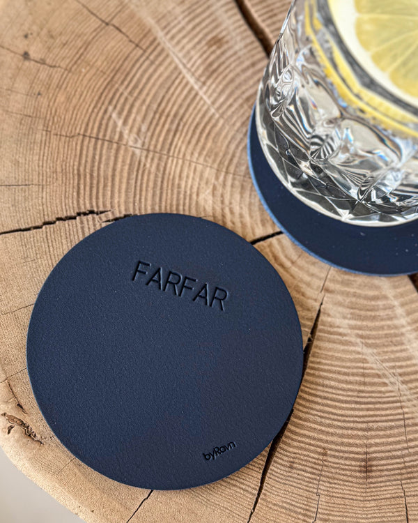 MOKKA round coaster in navy blue leather with the engraving "Farfar" on a wooden slab next to a crystal glass with lemon