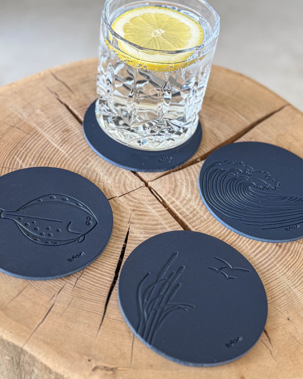 MOKKA summer house coasters in navy blue leather embossed with flatfish, wave and marram grass on a wooden slab by a glass with lemon