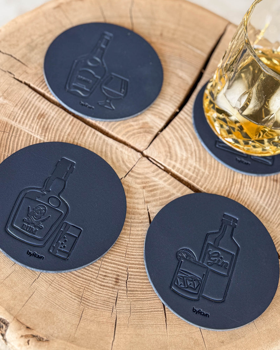 MOKKA spirits coasters in navy blue leather embossed with cognac, rum and gin on a wood slice by a crystal glass of whisky and ice