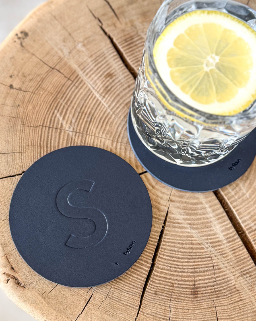 MOKKA coaster in navy blue leather embossed with the letter "S" on a wooden slab next to a crystal glass with lemon slice