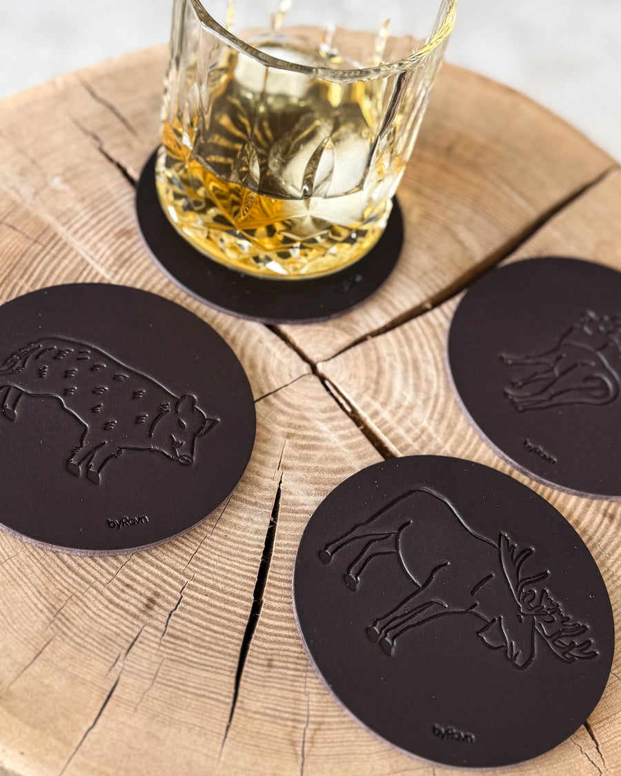 MOKKA hunting coasters in dark brown leather embossed with wild boar, elk and fallow deer on a wooden slab by a whisky glass