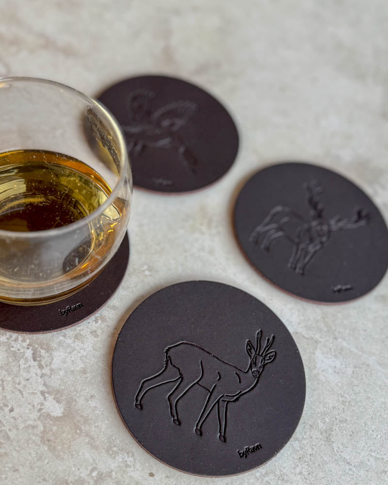 MOKKA hunting coasters in dark brown leather embossed with roebuck, mallard and red deer stag on a light table by a glass of whisky
