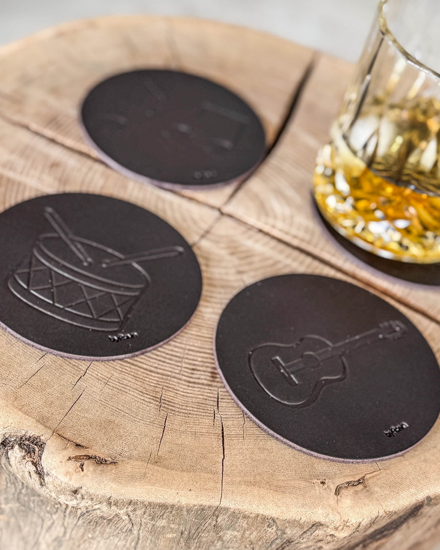 MOKKA music coasters in dark brown leather embossed with drum and guitar on a wood slice by a crystal glass of whisky
