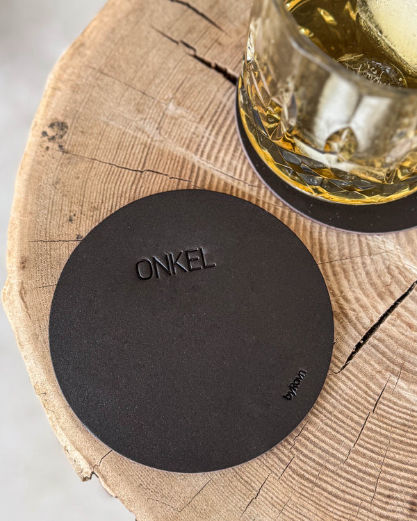 MOKKA round coaster in dark brown leather with the engraving "Onkel" on a wooden slab, whisky glass on a coaster behind