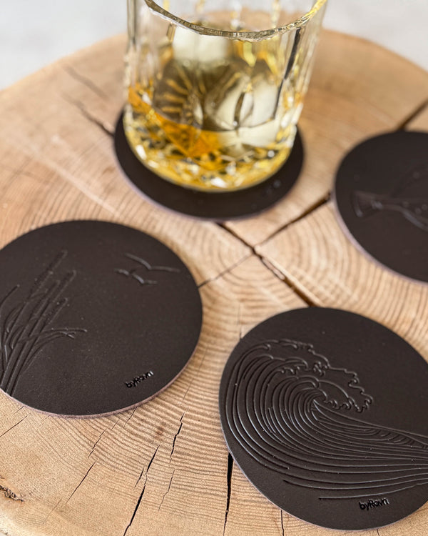 MOKKA summer house coasters in dark brown leather embossed with wave and marram grass with seagulls on a wooden slab by a whisky glass