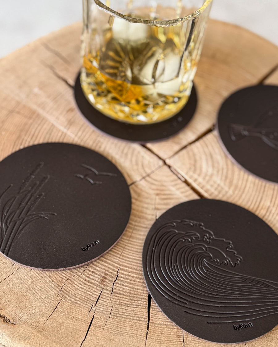 MOKKA summer house coasters in dark brown leather embossed with wave and marram grass with seagulls on a wooden slab by a whisky glass