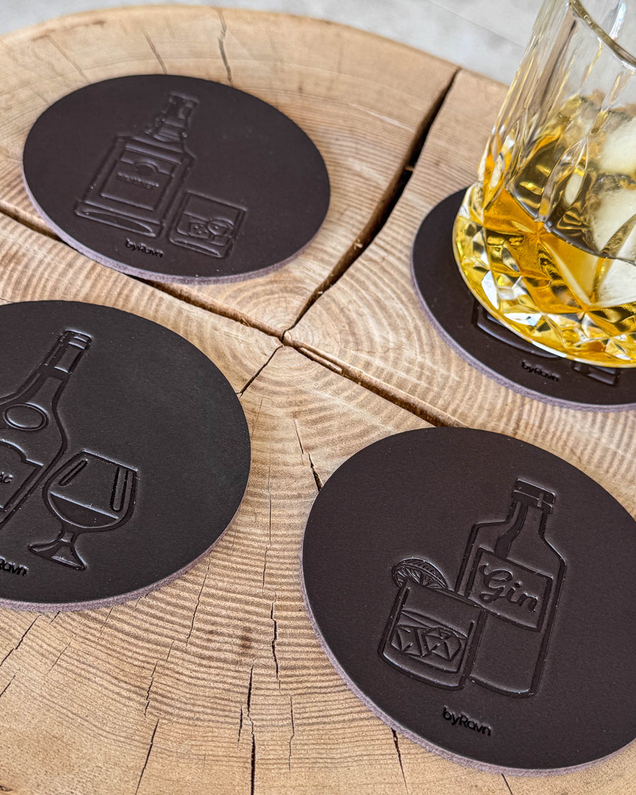 MOKKA spirits coasters in dark brown leather embossed with gin, cognac and whisky on a wooden slab by a glass of whisky with ice