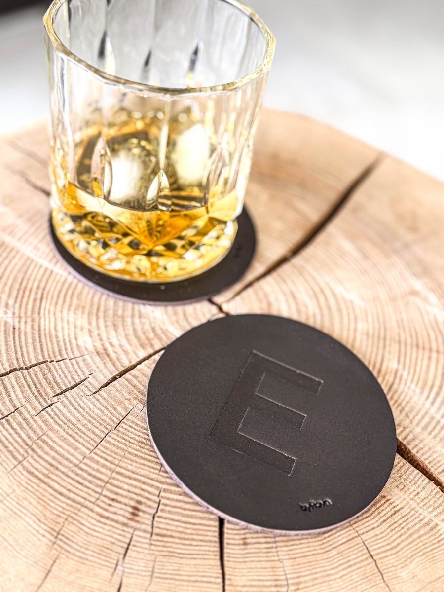 MOKKA coaster in dark brown leather embossed with the letter "E" on a wooden slab next to a whisky glass on a coaster