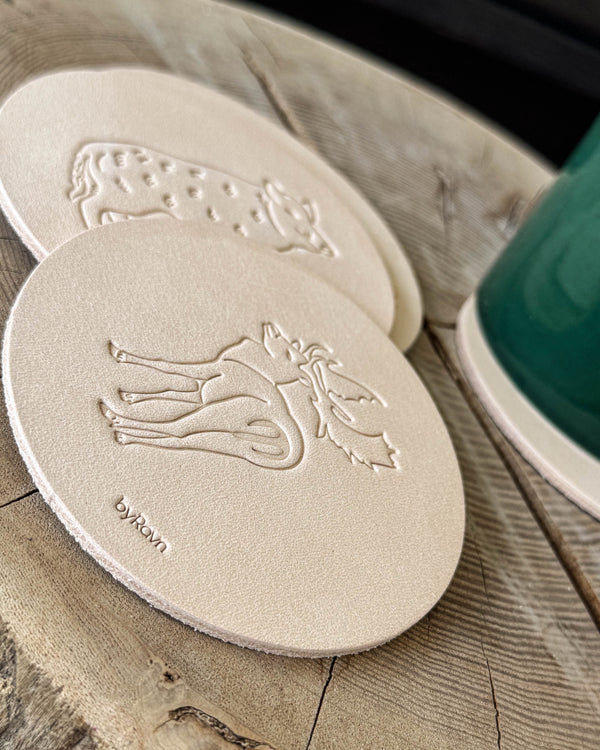 MOKKA hunting coasters in natural full-grain leather embossed with elk and wild boar on a wooden table next to a green cup