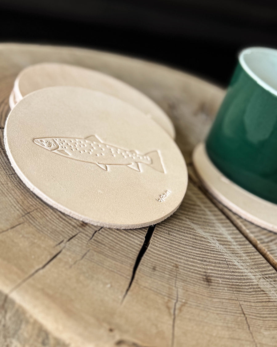 MOKKA angler coaster in natural full-grain leather embossed with a sea trout on a wooden table next to a green cup on a coaster