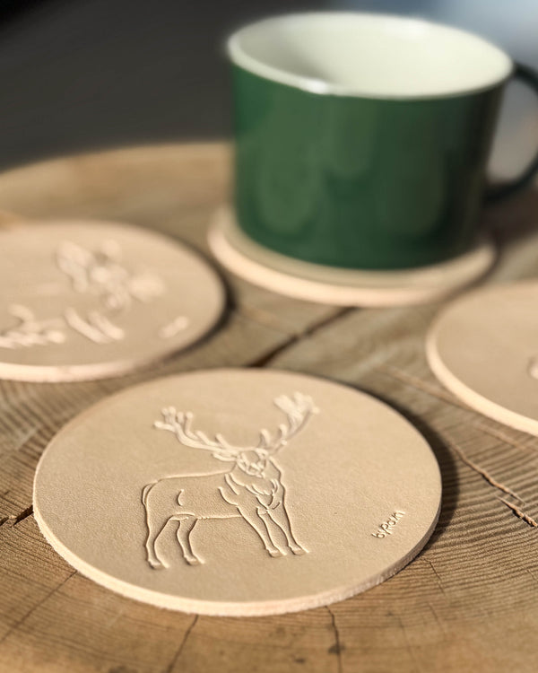 MOKKA hunting coaster in natural full-grain leather embossed with a red deer stag on a wooden table, more coasters and green cup behind
