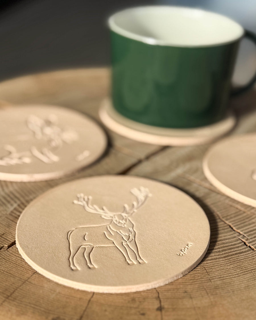 MOKKA hunting coaster in natural full-grain leather embossed with a red deer stag on a wooden table, more coasters and green cup behind