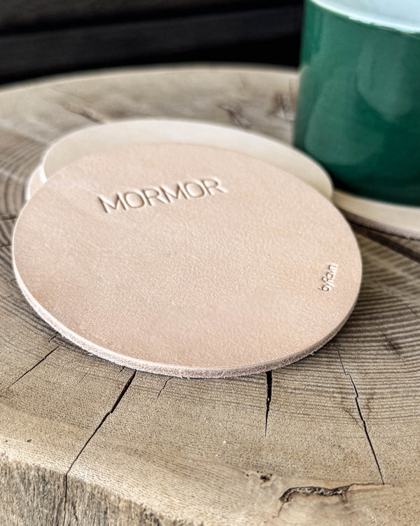 MOKKA round coasters in natural full-grain leather with the engraving "Mormor" on a wooden slab next to a green cup