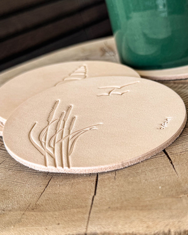 MOKKA summer house coasters in natural leather embossed with marram grass and seagulls, conch shell behind, on a wooden slab by a green cup