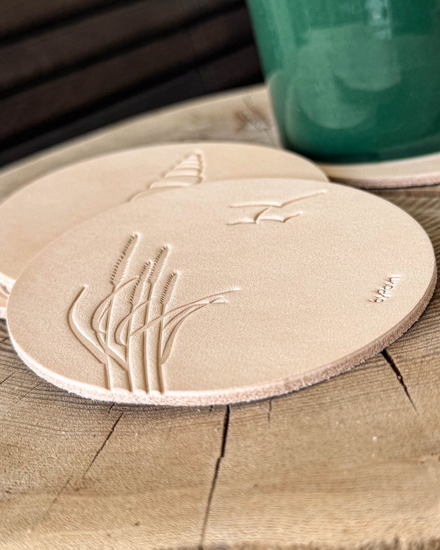 MOKKA summer house coasters in natural leather embossed with marram grass and seagulls, conch shell behind, on a wooden slab by a green cup
