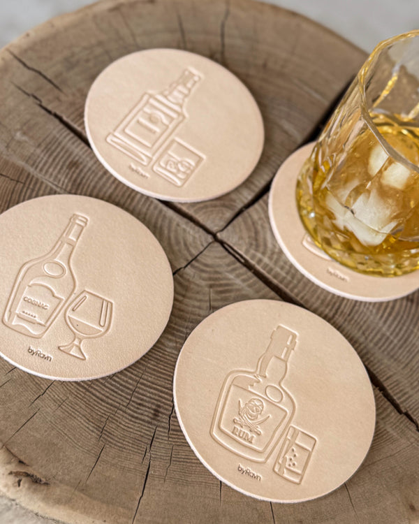 MOKKA spirits coasters in natural leather embossed with cognac, rum and whisky on a wooden slab by a glass of whisky with ice