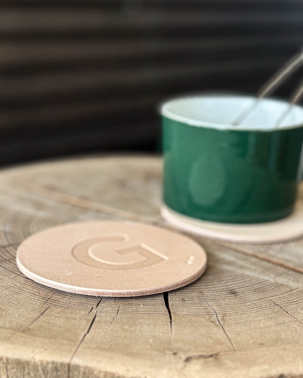 MOKKA coaster in natural leather embossed with the letter "G" on a wooden slab in front of a green cup with spoon