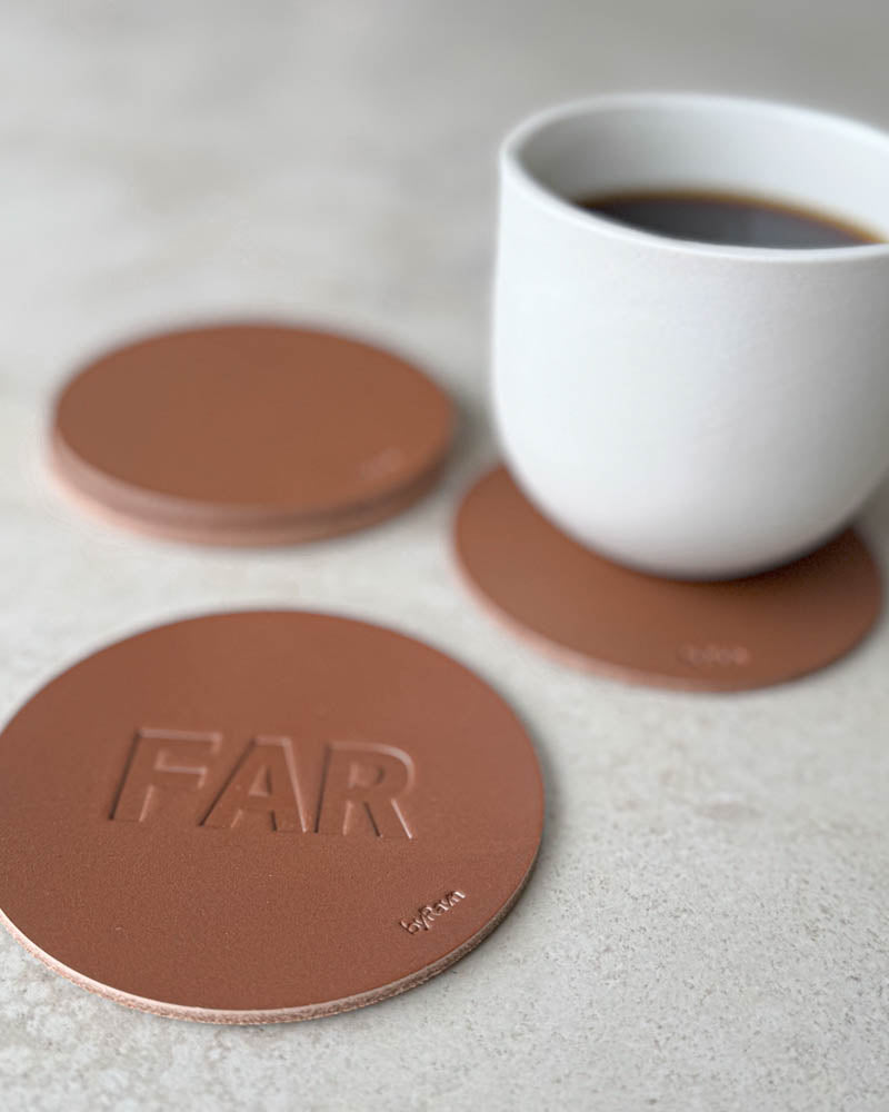 MOKKA coaster in cognac leather embossed "Far" in capital letters, more coasters and a white coffee cup behind