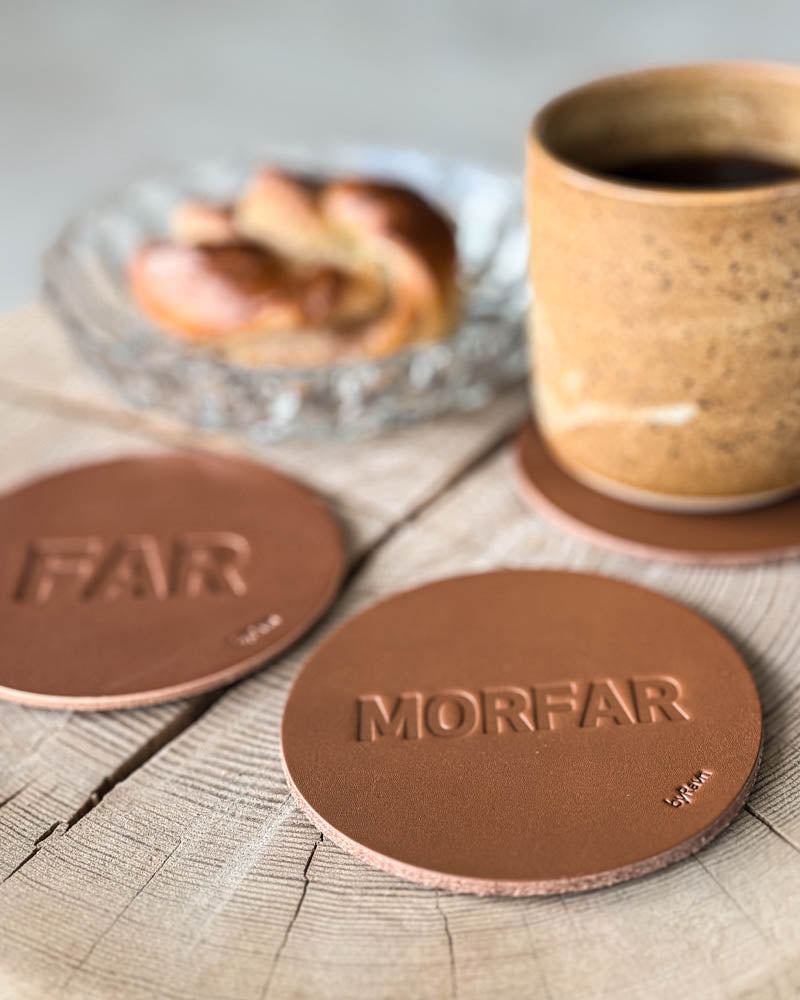 MOKKA coasters in cognac leather embossed "Far" and "Morfar" on a wooden slab with coffee cup and cinnamon bun on a glass plate