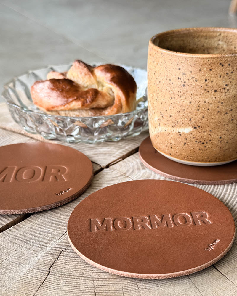 MOKKA coasters in cognac leather embossed "Mor" and "Mormor" on a wooden table with coffee cup and cinnamon bun in a glass bowl