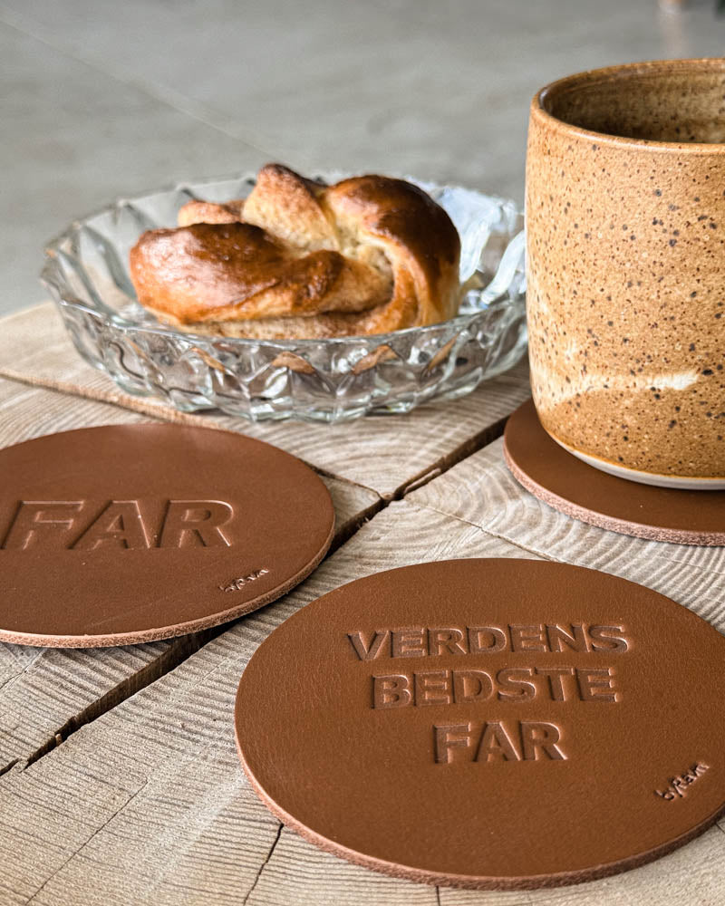 MOKKA coasters in cognac leather embossed "Far" and "Verdens bedste far" next to coffee cup and cinnamon bun on a wooden table
