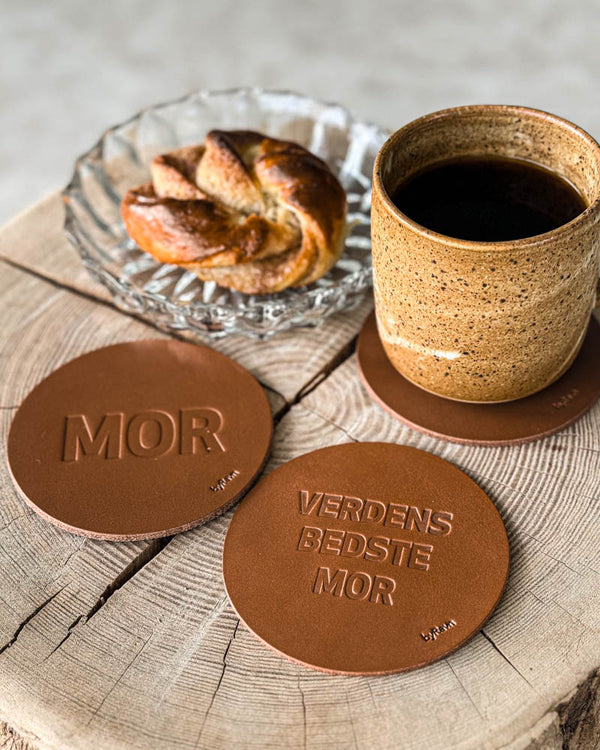 MOKKA coasters in cognac leather embossed "Mor" and "Verdens bedste mor" on a wooden slab with coffee cup and cinnamon bun