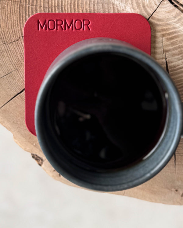 MOKKA square coaster in red leather with the engraving "Mormor" under a black coffee cup on a wooden slab, seen from above