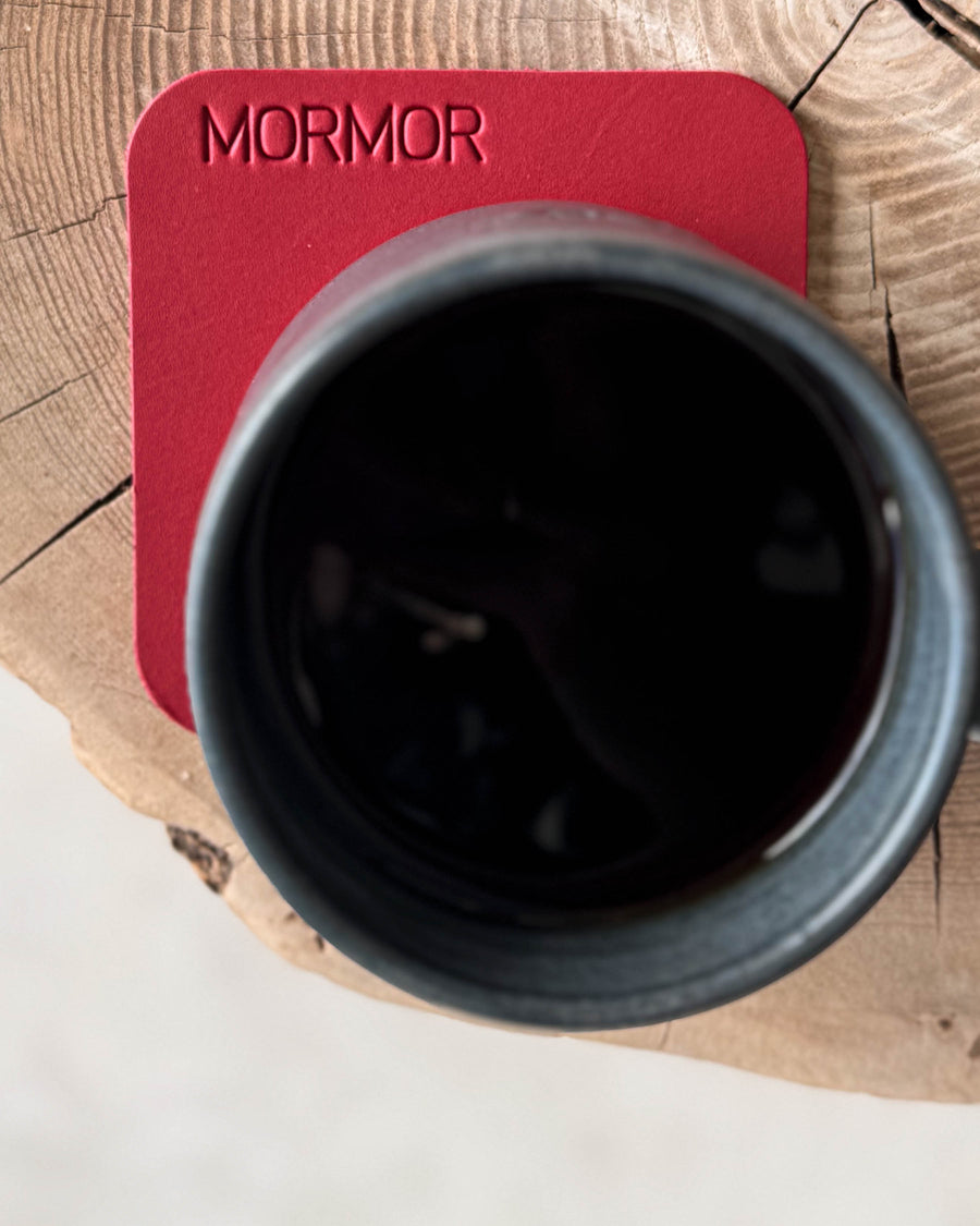 MOKKA square coaster in red leather with the engraving "Mormor" under a black coffee cup on a wooden slab, seen from above