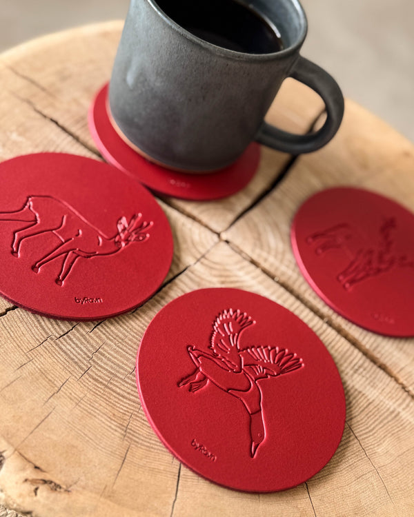 MOKKA hunting coasters in red leather embossed with flying mallard, roebuck and hare on a wooden slab next to a grey coffee cup