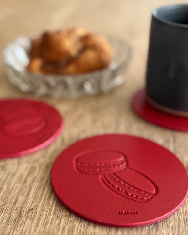 MOKKA coffee & cake coaster in red leather embossed with macarons on a wooden table by a black cup and cinnamon bun in a glass bowl