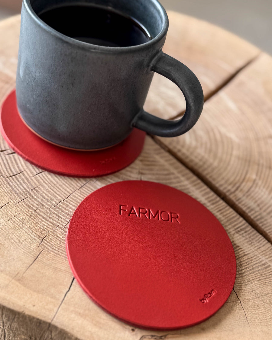 MOKKA round coaster in red leather with the engraving "Farmor" on a wooden slab, grey coffee cup on a coaster behind