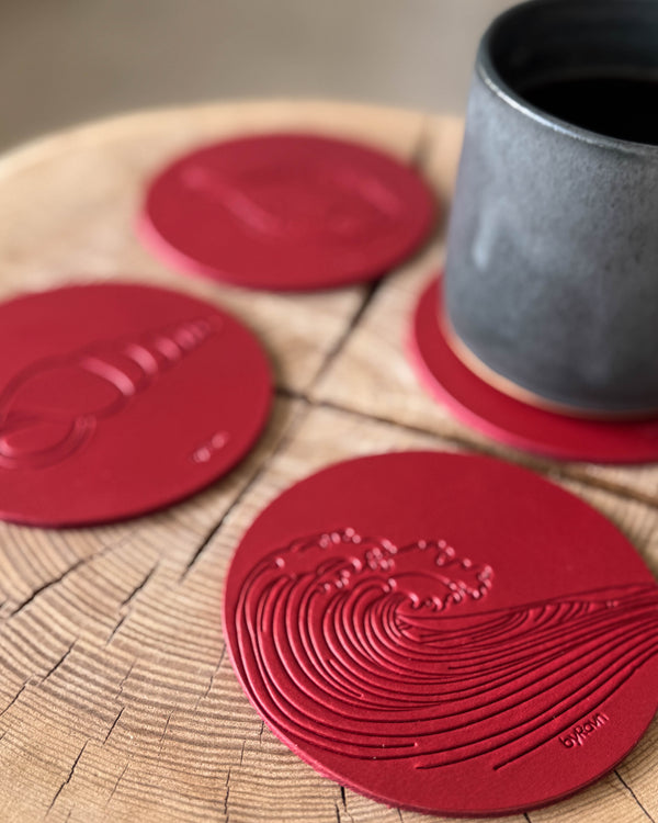 MOKKA summer house coasters in red leather embossed with wave and conch shell on a wooden slab next to a grey coffee cup on a coaster