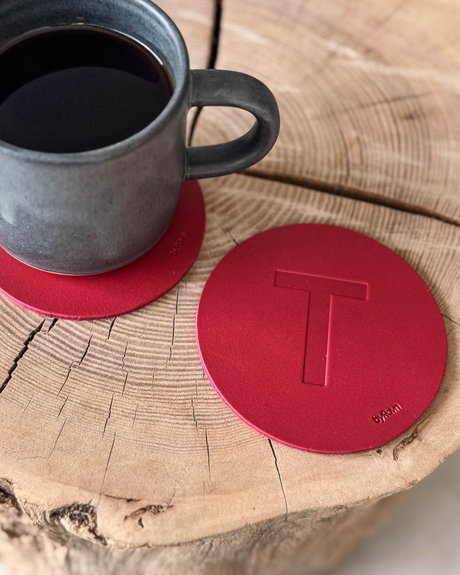 MOKKA coaster in red leather embossed with the letter "T" on a wooden slab next to a grey coffee cup on a coaster