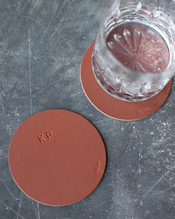 MOKKA round coasters in cognac leather engraved with the initials "K.P.", one under a crystal glass on a grey concrete table