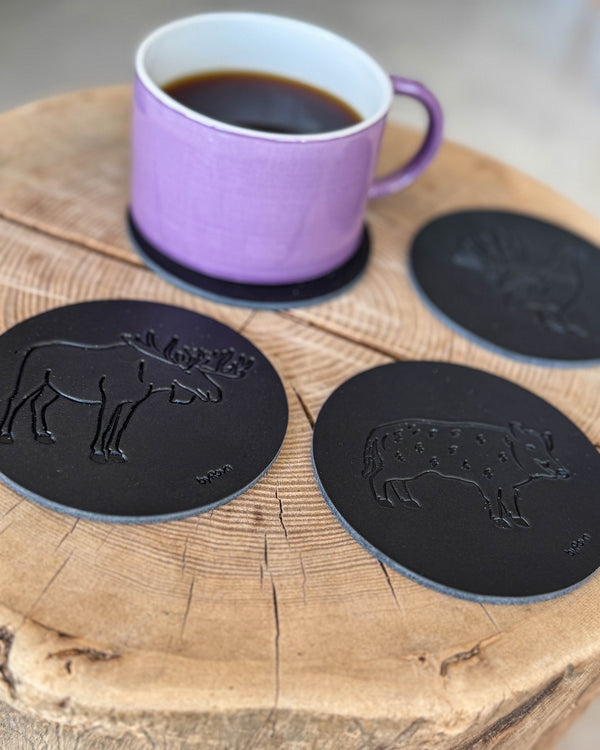 MOKKA hunting coasters in black leather embossed with elk, wild boar and capercaillie on a wooden slab with purple coffee cup on a coaster