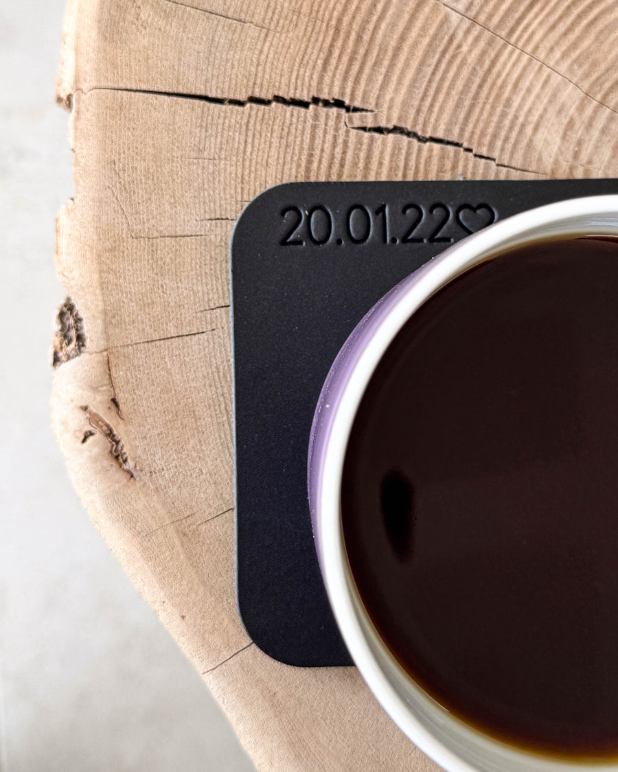 MOKKA square coaster in black leather with personalised engraving "20.01.22" ♡ under a coffee cup on a wooden slice