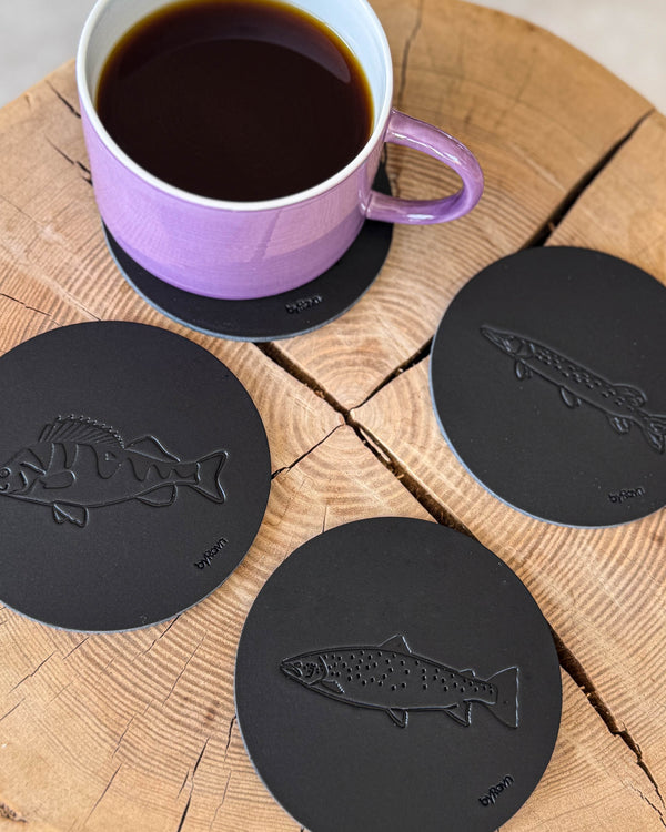 MOKKA angler coasters in black leather embossed with sea trout, perch and pike on a wooden slab next to a purple coffee cup