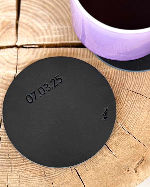MOKKA round coaster in black leather engraved with the date "07.03.25" on a wooden slab, purple coffee cup on a coaster behind