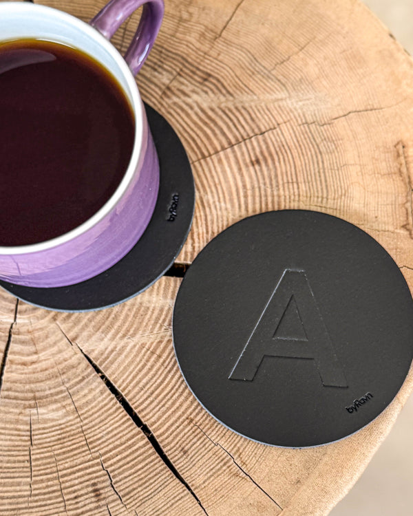 MOKKA coaster in black leather embossed with the letter "A" on a wooden slab next to a purple coffee cup on a coaster