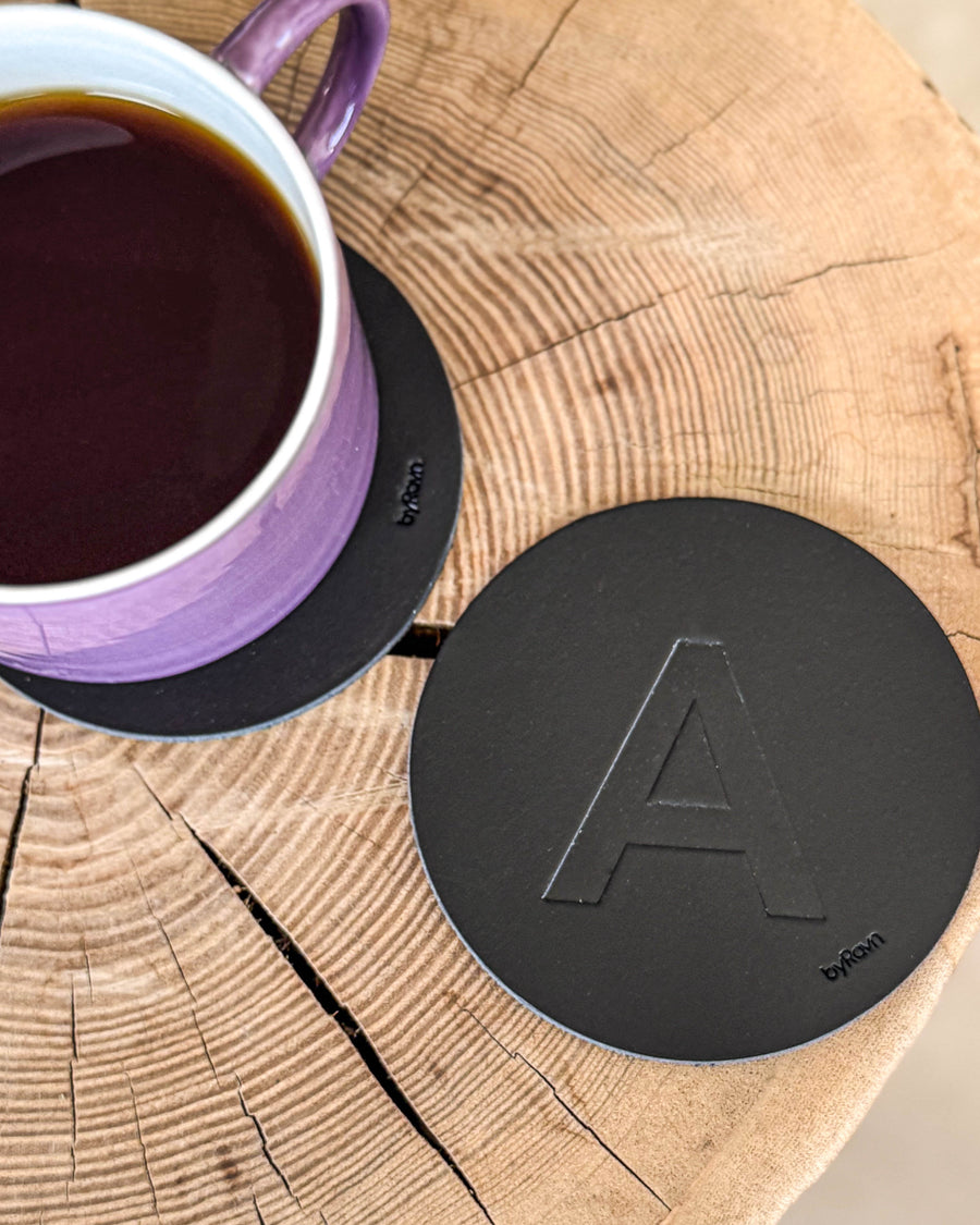 MOKKA coaster in black leather embossed with the letter "A" on a wooden slab next to a purple coffee cup on a coaster