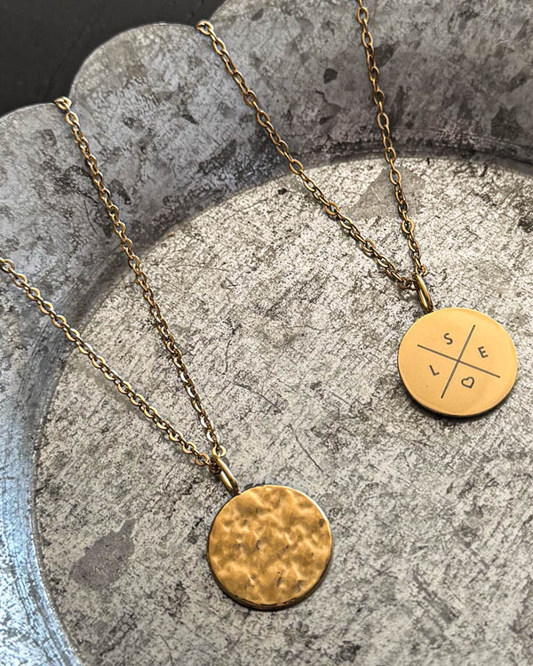 MOON X necklace in gold-plated stainless steel with cross and initials "S E L" and heart engraved, hammered coin alongside, on zinc tray